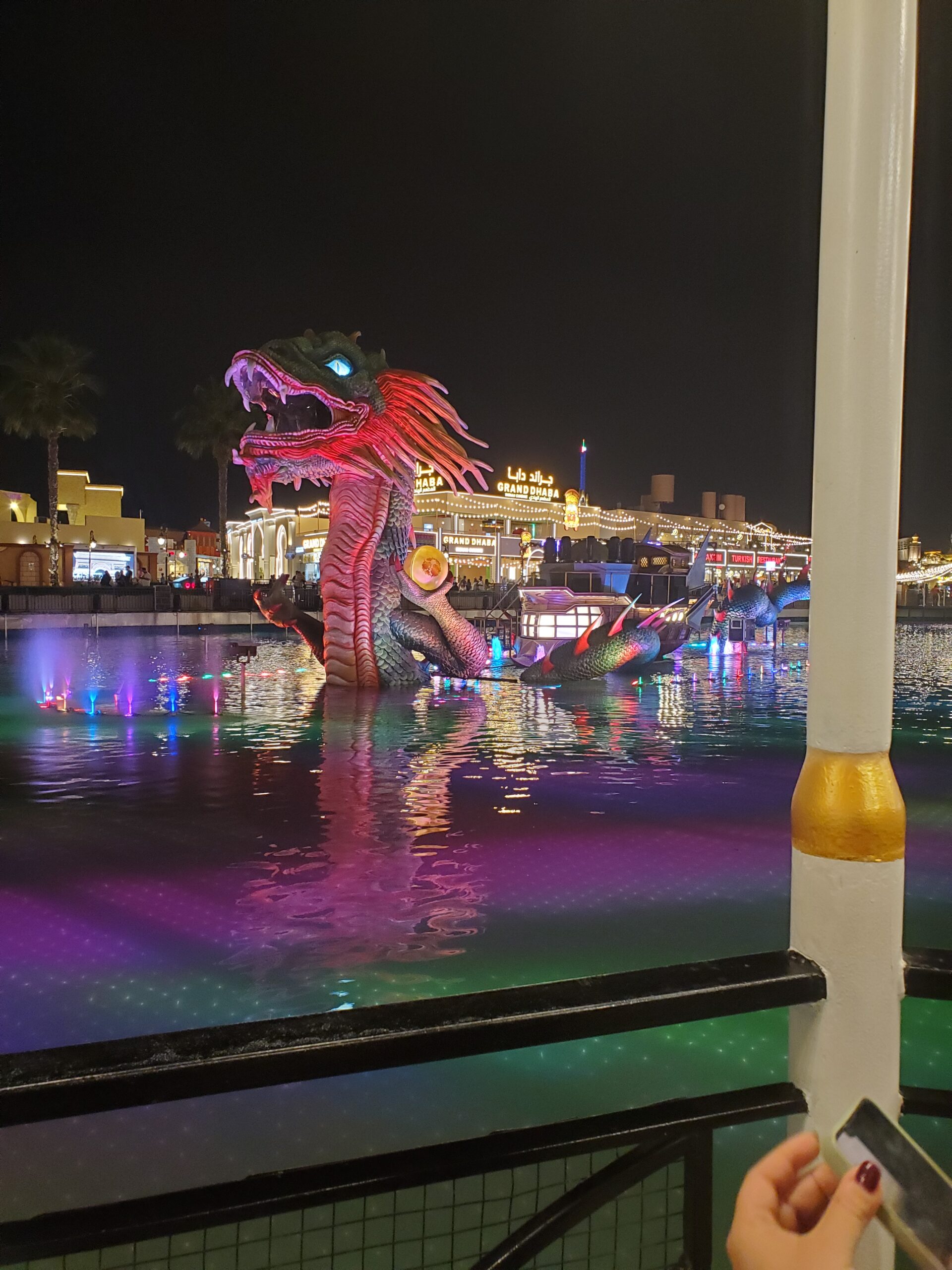 Night view of Dubai on the river with red and green dragon boat. Behind the river is a line of buildings lit up with lights 