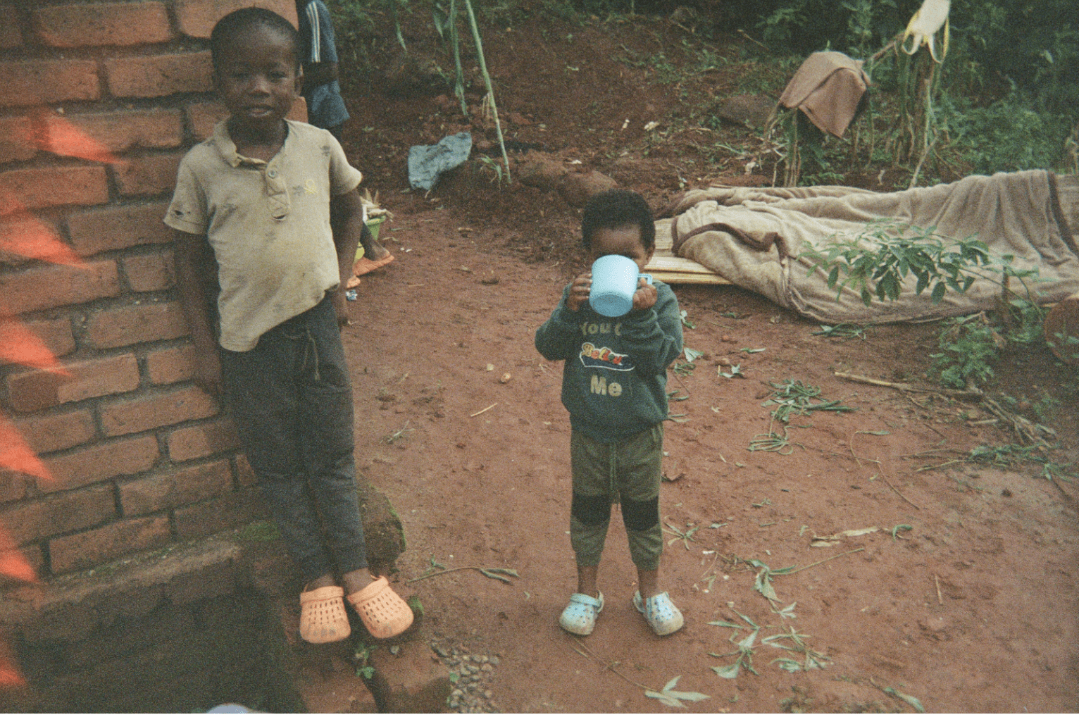 Two boys standing by a brick wall. The younger boy is drinking from a blue cup.