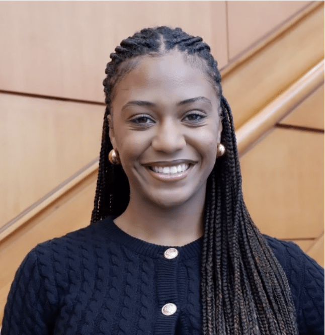 Jordan Lockwood stands with neatly braided hair and gold earrings is smiling warmly at the camera. She is dressed in a dark knitted sweater with gold buttons. The background features wooden paneling and a staircase, creating a professional and formal atmosphere.