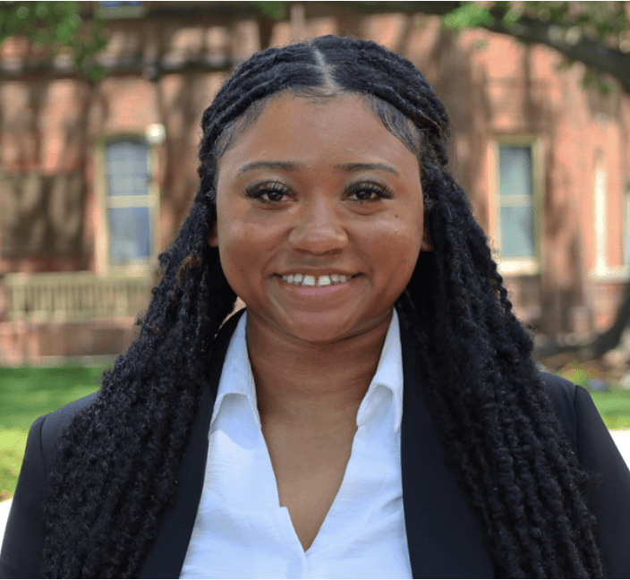 Alixandra Green stands outside in a sunny setting with green grass and a blurred background of a brick building. She has long, intricate braids framing her face and wears a confident smile. Her attire consists of a white collared shirt and a dark blazer, giving a professional appearance.