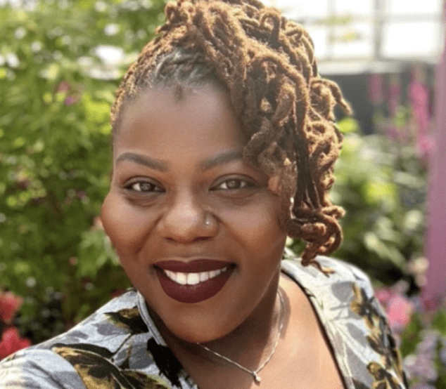 Dr. Vanessa Goodar stands with a warm smile in a garden, surrounded by lush greenery and vibrant flowers. Her hair is styled in long, intricately braided locks, pulled up and to the side. She wears a floral-patterned top that complements the natural setting. The sunlight illuminates her face, highlighting her joyful expression and the garden's colorful blooms.